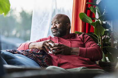 A mature multiracial man at home, sitting on a couch near a window, holding a cup of tea. He is leaning back, looking away, thinking, with a serious but relaxed expression.