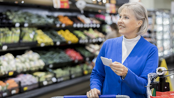A cheerful senior woman looks up at an abundant wall of produce at the grocery store. She is pushing a cart and holding a shopping list.
